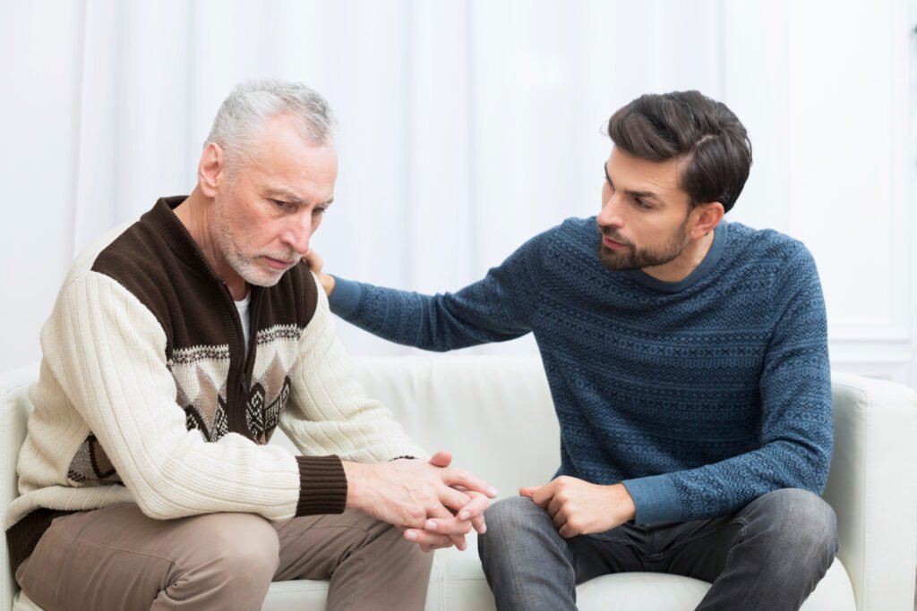 Adult son placing a comforting hand on his withdrawn, downcast father's shoulder while sitting beside him on a sofa
